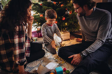 Ein Junge öffnet sein Weihnachtsgeschenk, eine Familie sitzt am Boden vor dem Weihnachtsbaum und packt Weihnachtsgeschenke aus. - JLPSF10912
