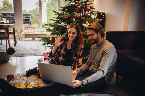 Ein Paar benutzt einen Laptop für einen Videoanruf mit der Familie an einem Weihnachtstag. Ein Mann und eine Frau führen einen Videoanruf mit einem geschmückten Weihnachtsbaum im Hintergrund. - JLPSF10918