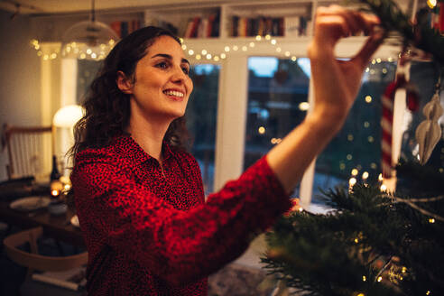 Frau beim Schmücken des Weihnachtsbaums zu Hause. Frau hängt Dekorationsartikel an den Weihnachtsbaum im Haus. - JLPSF10941