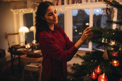 Frau schmückt Weihnachtsbaum allein zu Hause. Frau hängt Ornamente an einen Weihnachtsbaum. - JLPSF10943