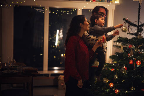 Kleine Familie schmückt gemeinsam den Weihnachtsbaum zu Hause. Eltern mit Kind schmücken das Haus für die Weihnachtsfeier. - JLPSF10945