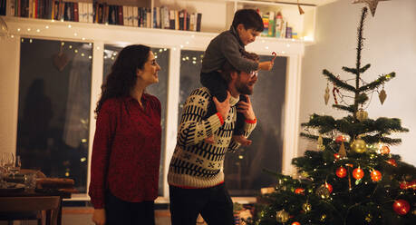 Mann mit seinem Sohn auf den Schultern, der ihm beim Schmücken des Weihnachtsbaums hilft; Innenaufnahme eines weihnachtlich geschmückten Familienhauses. - JLPSF10946