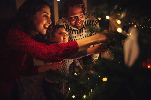 Eine dreiköpfige Familie bereitet einen Tannenbaum für die Weihnachtsfeier zu Hause vor. Eine Familie schmückt gemeinsam zu Hause einen Weihnachtsbaum. - JLPSF10948