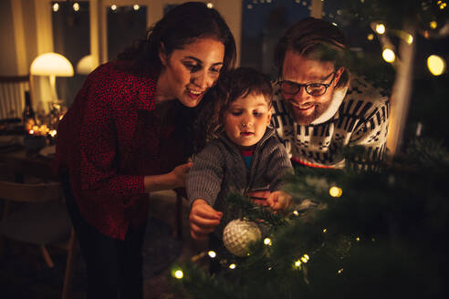 Junge bindet dekorative Kugeln an Weihnachtsbaum mit Eltern zu Hause. Weihnachtsferien Vorbereitungen in skandinavischen Haus. - JLPSF10949