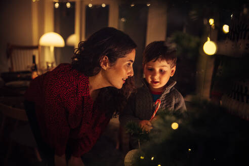 Mutter und Sohn legen eine Zuckerstange auf den Weihnachtsbaum zu Hause. Junge Frau im Gespräch mit ihrem Sohn beim Schmücken des Weihnachtsbaums. - JLPSF10950