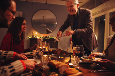 Elderly man serving food to his family sitting at dinner table on Christmas eve. Family having Christmas dinner together in warn and cozy scandinavian home. - JLPSF10962