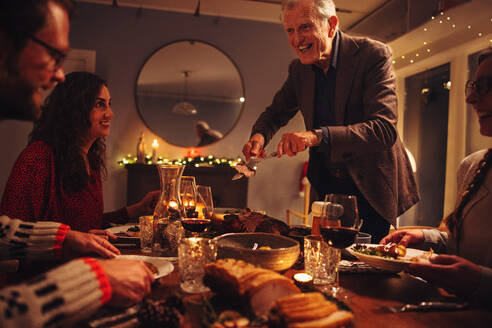 Älterer Mann, der seiner Familie am Weihnachtsabend am Tisch sitzend das Essen serviert. Familie beim gemeinsamen Weihnachtsessen in einem warmen und gemütlichen skandinavischen Haus. - JLPSF10962