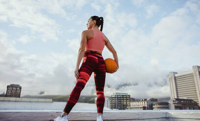 Rear view of female in sportswear holding basketball on rooftop. Sportswoman standing outdoors with basketball in hand. - JLPSF10980