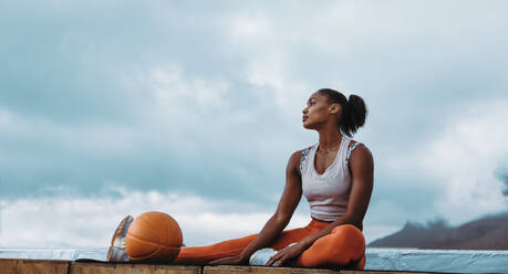 Fitness-Frau entspannt sich nach dem Training auf dem Dach. Sportliche Frau mit Medizinball und sitzt im Freien und schaut weg. - JLPSF11000