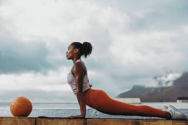 Seitenansicht einer Frau mit fitten Körper tun Rücken Stretching-Übung im Freien. Fitness-Frau übt Kobra Pose Yoga-Workout auf dem Dach mit einem Basketball vor. - JLPSF11001