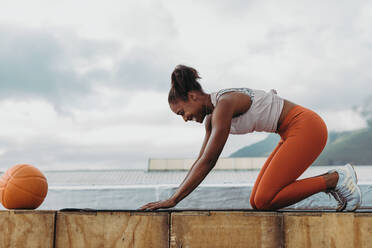Seitenansicht einer lächelnden Frau beim Workout im Freien mit einem Basketball vor sich. Sportlerin übt Yoga auf dem Dach. - JLPSF11003
