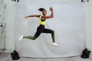 Sportlerin im Trainingsanzug springt hoch in der Luft auf einem Dach. Afrikanische Sportlerin, die im Freien läuft und Ausfallschritte macht. - JLPSF11023