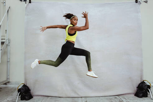Sportswoman in exercise outfit jumping high in midair on rooftop. African athlete woman doing running and lunge working outdoors. - JLPSF11023