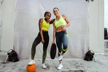 Sportlerin mit Fuß auf Basketball stehend mit ihrer Freundin, die eine Hantelscheibe hält und in die Kamera schaut, auf einem Dach. Frauen in Sportkleidung posieren nach dem Training. - JLPSF11060