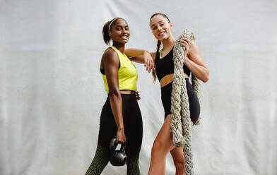 Fit woman with kettlebell and her friend holding battle rope. Females in sportswear standing with gym equipment on white background. - JLPSF11070