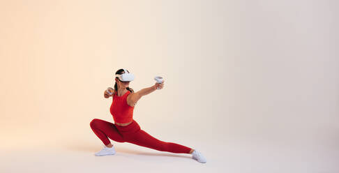 Active young woman working out in virtual reality. Sporty young woman doing fitness exercises using a pair of gaming controllers. Athletic young woman wearing a virtual reality headset in a studio. - JLPSF11075