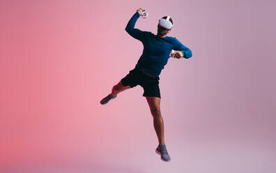 Energiegeladener junger Mann, der einen virtuellen Ball mit Hilfe von Gaming-Controllern wirft. Aktiver junger Mann, der mitten in der Luft springt, während er eine Virtual-Reality-Brille trägt. Junger Mann, der ein virtuelles Handballspiel spielt. - JLPSF11110