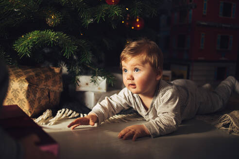 Niedlicher kleiner Junge, der zu Hause unter dem Weihnachtsbaum liegt und die Weihnachtsgeschenke betrachtet. - JLPSF11114