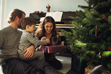 Junge öffnet sein Weihnachtsgeschenk mit Eltern sitzend. Kleine Familie sitzt in der Nähe des Weihnachtsbaums und packt Geschenke aus. - JLPSF11115