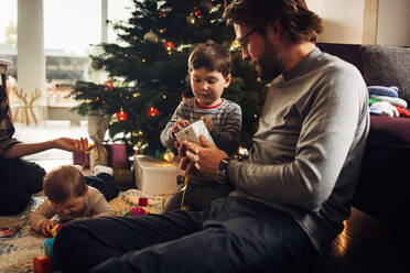 Vater und Sohn beim Auspacken der Weihnachtsgeschenke, Familie sitzt im Wohnzimmer vor dem Weihnachtsbaum und packt Geschenke aus. - JLPSF11117