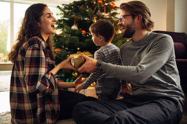 Frau, die ein Geschenk für Weihnachten und Neujahr von ihrem Mann erhält. Glückliche junge Familie, die am Weihnachtsabend am Weihnachtsbaum sitzt. - JLPSF11120