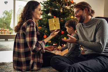 Ehepaar sitzt mit ihrem Sohn am Weihnachtsbaum und tauscht zu Hause Geschenke aus. Schöne kleine Familie, die Weihnachten zu Hause feiert. - JLPSF11121
