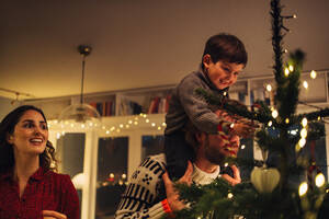 Junge sitzt auf der Schulter seines Vaters beim Schmücken des Weihnachtsbaums. Familie schmückt gemeinsam den Weihnachtsbaum zu Hause. - JLPSF11129