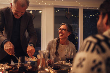 Älterer Mann, der seiner Familie am Weihnachtsabend das Essen serviert. Familie beim gemeinsamen Weihnachtsessen. - JLPSF11130