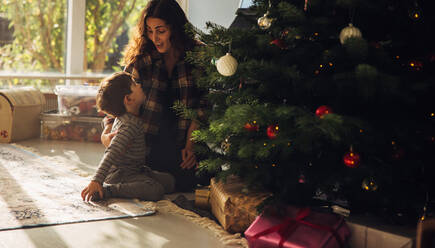 Mutter mit ihrem Kind feiert Weihnachten. Frau mit ihrem Sohn sitzt neben einem geschmückten Weihnachtsbaum mit vielen Geschenkboxen; - JLPSF11140
