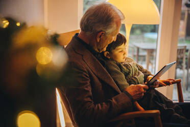 Älterer Mann mit ihrem Enkel, der einen Videoanruf auf einem digitalen Tablet führt. Großvater und Enkel sitzen auf einem Stuhl und benutzen ein digitales Tablet während Weihnachten. - JLPSF11152