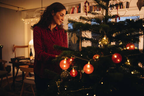 Frau schmückt Weihnachtsbaum allein zu Hause. Frau hängt Ornamente an einen Weihnachtsbaum in der Familie zu Hause. - JLPSF11158