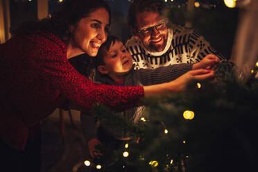 Kleine Familie beim gemeinsamen Schmücken des Weihnachtsbaums, Eltern mit Kind beim Schmücken zu Hause. - JLPSF11160
