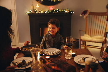 Ein Junge sitzt am Weihnachtsabend am Esstisch und schaut seine Mutter an. Ein Kind beim Weihnachtsessen mit seiner Familie. - JLPSF11162