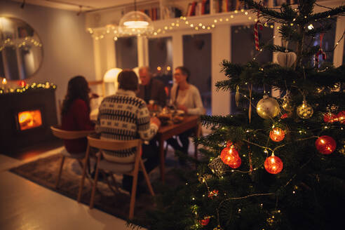 Innenaufnahme eines warmen und gemütlichen skandinavischen Hauses, das für das Weihnachtsfest geschmückt ist. Der Fokus liegt auf dem geschmückten Weihnachtsbaum mit der Familie, die am Esstisch sitzt und ein Weihnachtsabendessen im Hintergrund hat. - JLPSF11165