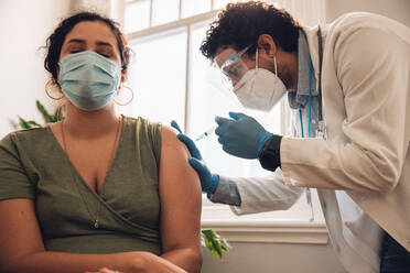 Woman getting a vaccine injection from doctor at home. Female wearing protective face mask receiving covid-19 vaccine by a healthcare professional at her home. - JLPSF11176