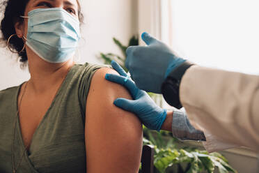 Female looking at healthcare professional while getting vaccine injection on her arm. Woman in face mask getting covid vaccine at home. - JLPSF11178