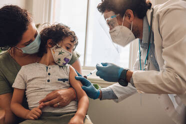 Arzt gibt einem Kind, das mit seiner Mutter zu Hause sitzt, eine Covid-19-Impfung. Junge mit Gesichtsmaske, der zu Hause von medizinischem Fachpersonal geimpft wird. - JLPSF11181