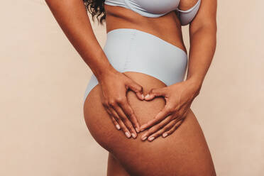 Self loving woman making a heart shape on her thigh in a studio. Closeup of an anonymous woman standing in blue underwear. Young woman embracing her natural body against a studio background. - JLPSF11193