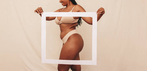 Happy young woman holding a picture frame in front of her body in a studio. Confident young woman feeling comfortable in her own body. Young woman wearing beige underwear against a studio background. - JLPSF11200