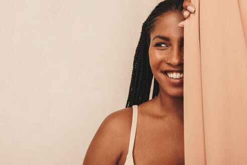 Carefree young woman smiling while standing behind a studio curtain. Confident young woman wearing a beige bra against a studio background. Body positive young woman embracing her natural beauty. - JLPSF11206