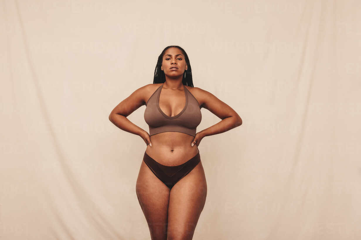 Self-confident young woman standing in brown underwear. Body positive young  woman looking at the camera while standing against a studio background. Young  woman embracing her natural body and curves. stock photo