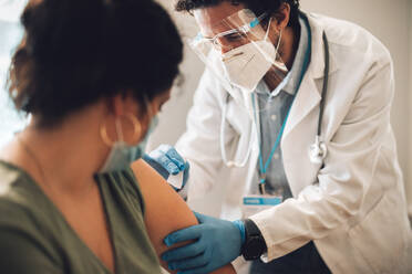 Healthcare professional giving vaccine to female at her home. Doctor wearing protective personal equipment giving coronavirus vaccination to a woman. - JLPSF11219