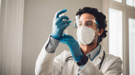 Doctor wearing face mask and face shield filling injection syringe from vial. Healthcare professional in protective workwear preparing coronavirus vaccine injection. - JLPSF11222