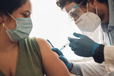 Close-up of a woman receiving covid-19 vaccine at home. Healthcare worker vaccinating a female in face mask at home. - JLPSF11226