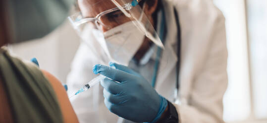 Close-up of a medical professional in protective worker wear doing vaccination. Doctor giving coronavirus vaccine during home visit. - JLPSF11227