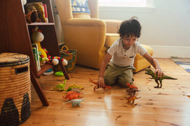 Fantasievoller kleiner Junge, der mit buntem Dinosaurierspielzeug in seinem Spielbereich zu Hause spielt. Liebenswerter kleiner Junge, der auf dem Boden kniet und ein Kampfspiel mit Tierspielzeug spielt. - JLPSF11236