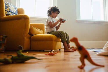 Kleiner Junge spielt mit buntem Tierspielzeug in seinem Spielbereich zu Hause. Liebenswerter kleiner Junge sitzt auf einer Couch und spielt ein Kampfspiel mit Dinosaurier-Spielzeug. - JLPSF11238