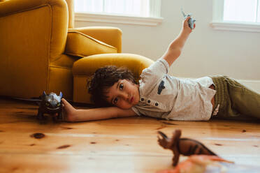 Niedlicher Junge, der zu Hause mit seinem Spielzeug spielt. Ethnischer Junge, der mit Dinosaurier-Spielzeug spielt, während er auf dem Boden in seinem Spielbereich liegt. - JLPSF11239