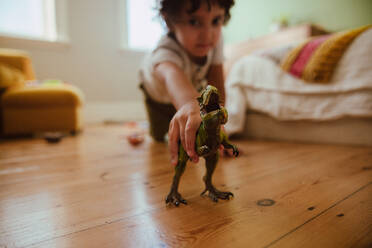 Ethnischer Junge, der zu Hause mit einem T-Rex-Dinosaurier spielt. Liebenswerter kleiner Junge, der ein brüllendes Dinosaurier-Spielzeug hält, während er sich in seinem Spielbereich hinkniet. - JLPSF11242