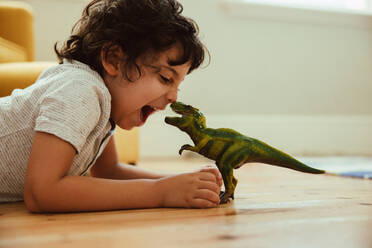 Adventurous young boy imitating a dinosaur toy while lying on the floor in his play area. Creative little boy having fun during playtime at home. - JLPSF11245